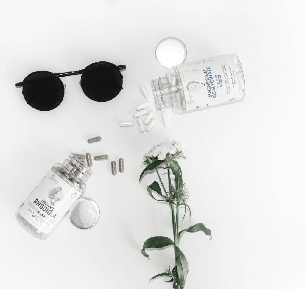Flatlay of magnesium and rhodiola supplement bottles with scattered capsules, black sunglasses, and a flower on a white surface.